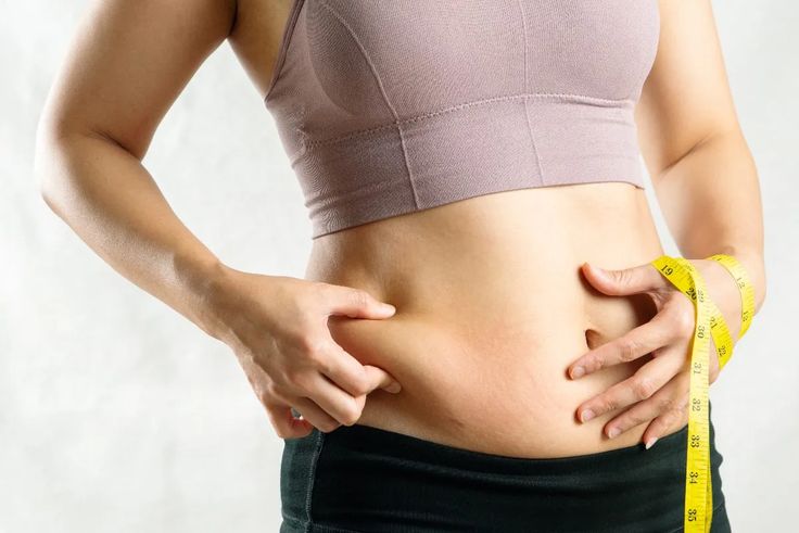 Woman holding stubborn abdominal fat while measuring her waist, representing liposuction in India for body contouring after weight loss.